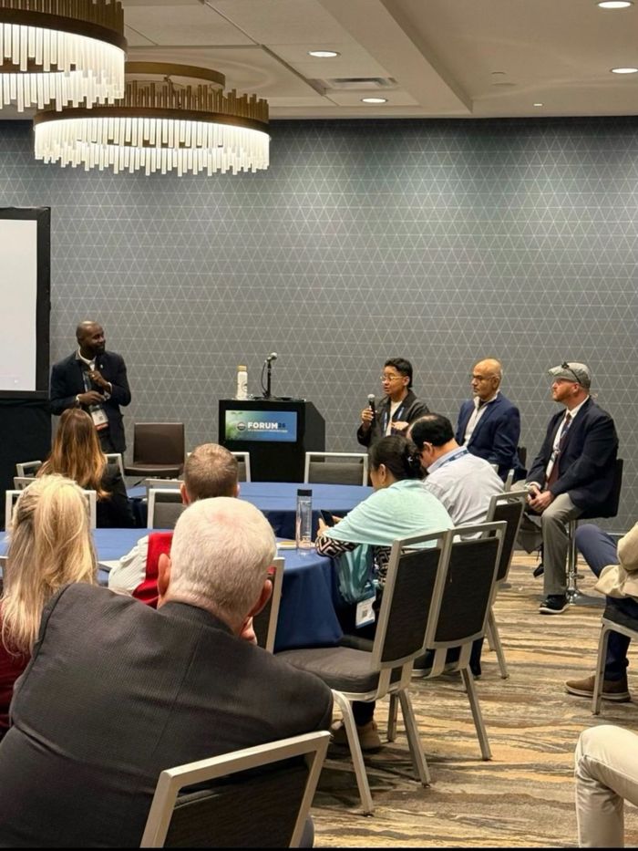 A panel discussion at a conference, with one panelist speaking into a microphone while the others listen